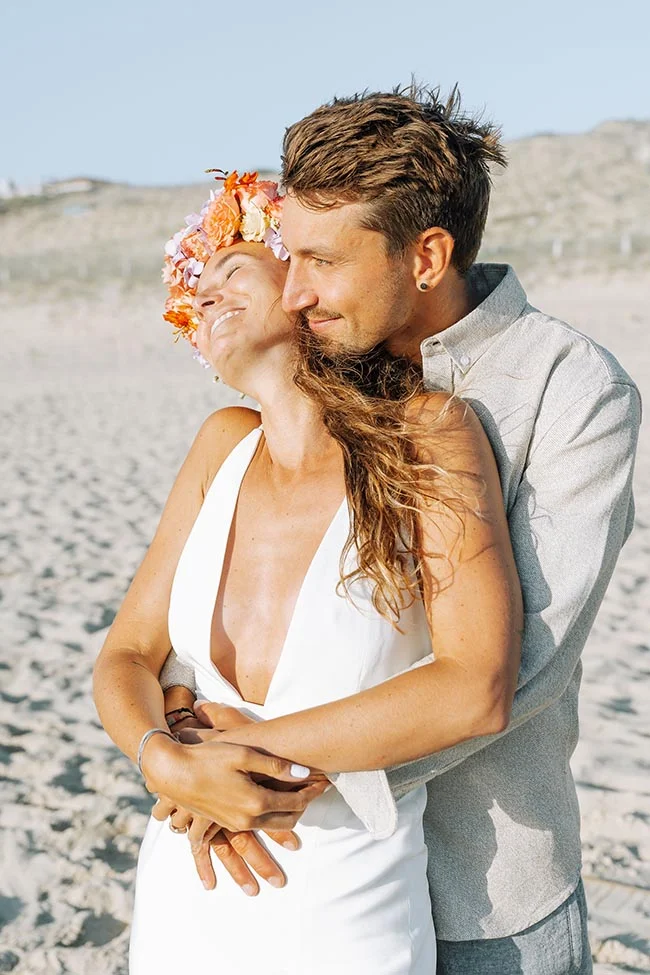 Couple marié s'enlaçant tendrement sur une plage de sable blanc, la mariée portant une couronne de fleurs colorées.