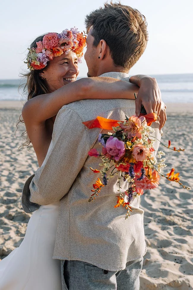 Jeunes mariés s'enlaçant sur une plage au coucher du soleil, la mariée portant une couronne de fleurs colorées.