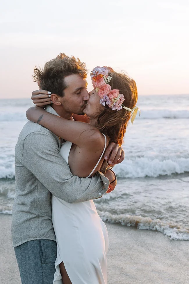 Jeune couple s'embrassant sur une plage au coucher du soleil, la mariée portant une couronne de fleurs colorées.