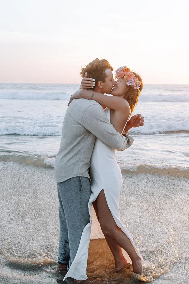 Jeunes mariés s'embrassant sur une plage au coucher du soleil, elle portant une couronne de fleurs et une robe blanche.
