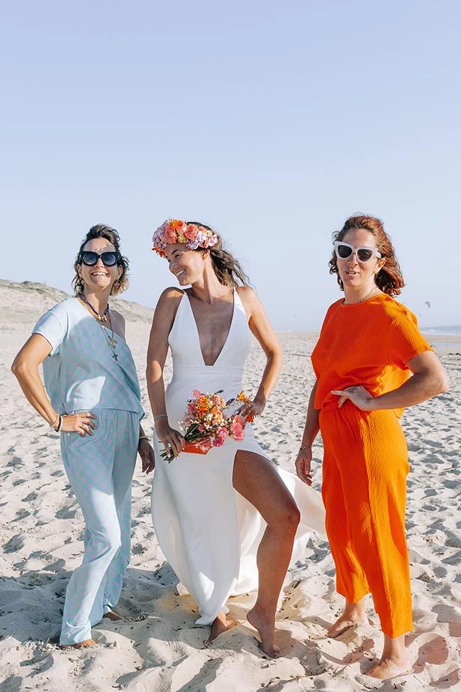 Trois femmes souriantes sur une plage de sable blanc, dont une mariée en robe blanche avec une couronne de fleurs colorées.