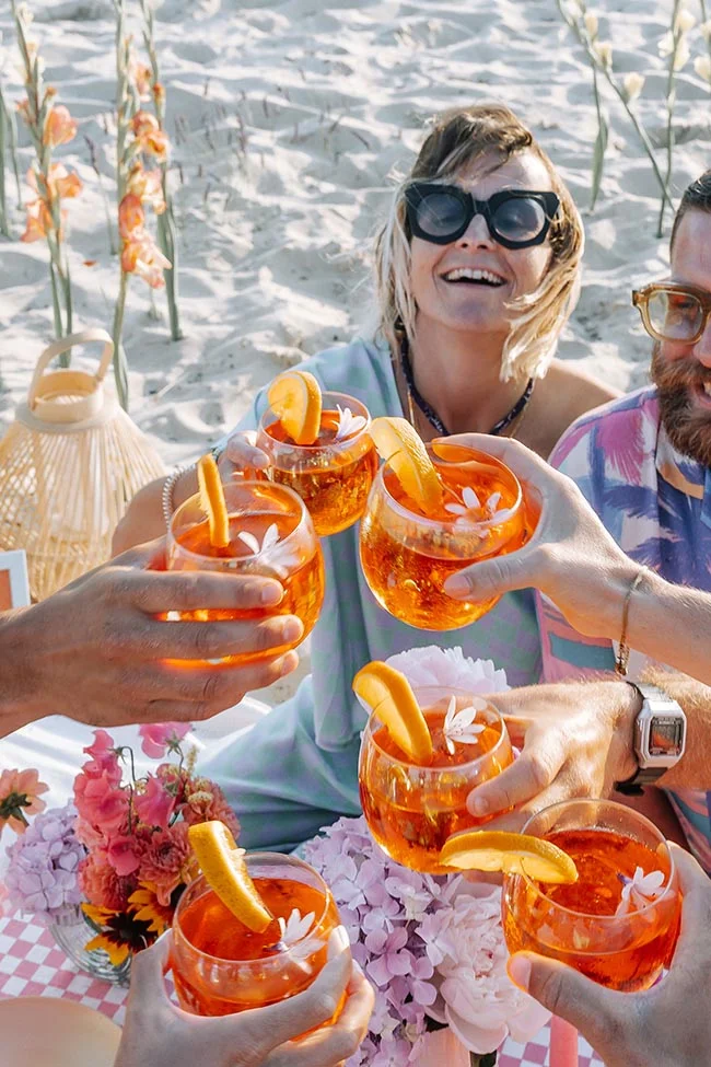 Des invités souriants trinquant avec des cocktails orangés à la plage dans une ambiance festive et lumineuse.
