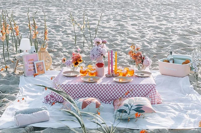 Pique-nique romantique et chic sur le sable avec nappe à carreaux rose, fleurs et champagne pour un mariage à la plage.