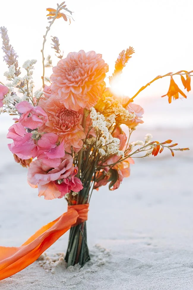 Bouquet de fleurs roses et orangées avec ruban satiné, posé sur le sable blanc sous une lumière solaire chaleureuse.