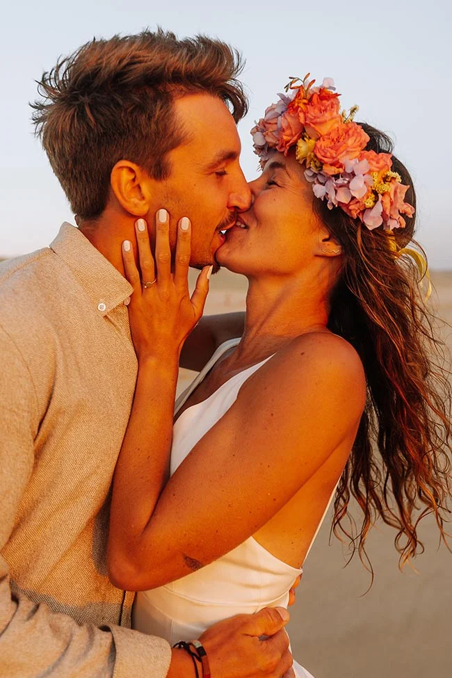 Jeunes mariés s'embrassant avec tendresse sur une plage au coucher du soleil, la mariée portant une couronne de fleurs.