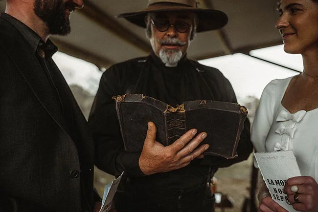 Trois personnes en tenues style western, dont un officiant lisant un vieux livre, dans une ambiance sobre et vintage.