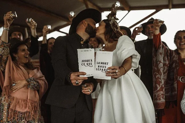 Jeunes mariés s'embrassant avec des pancartes, entourés d'invités festifs dans un style western aux tons chauds.