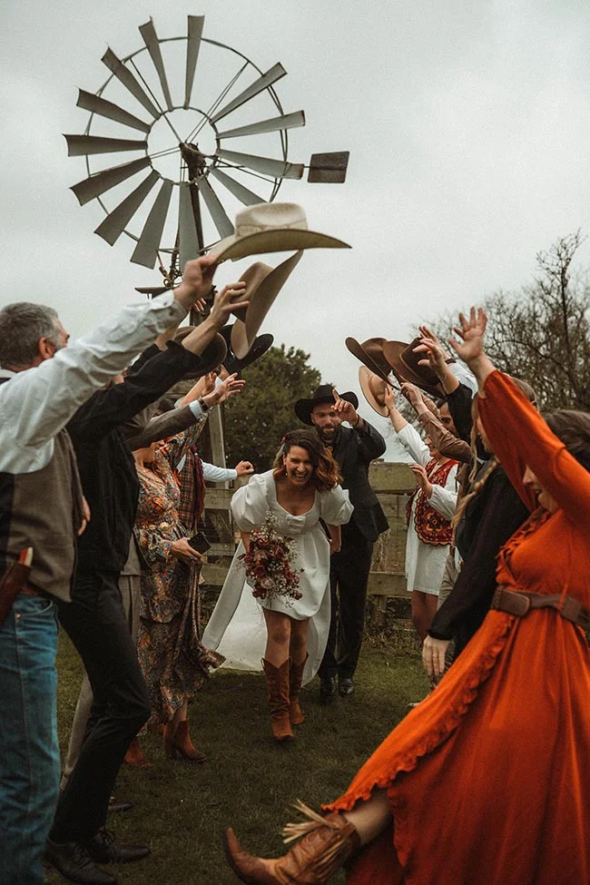 Jeunes mariés et invités célébrant avec joie un mariage western, lançant des chapeaux de cowboy dans un décor champêtre.