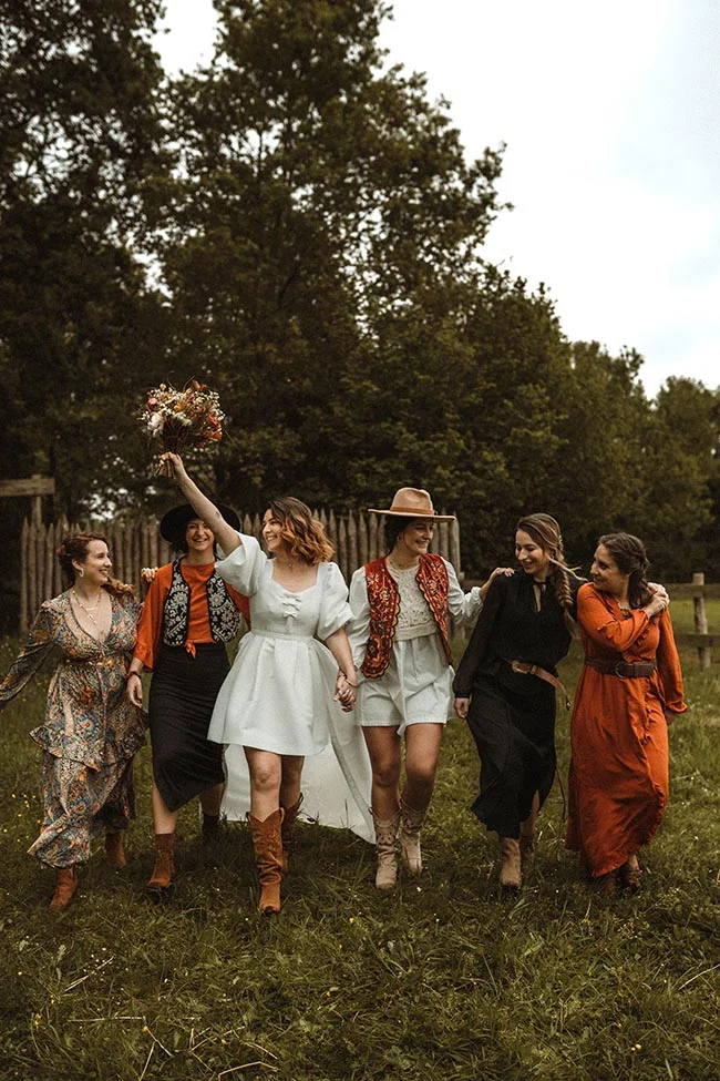 Cinq femmes souriantes en tenues style western et bohème marchent dans un champ, menées par la mariée en robe blanche.