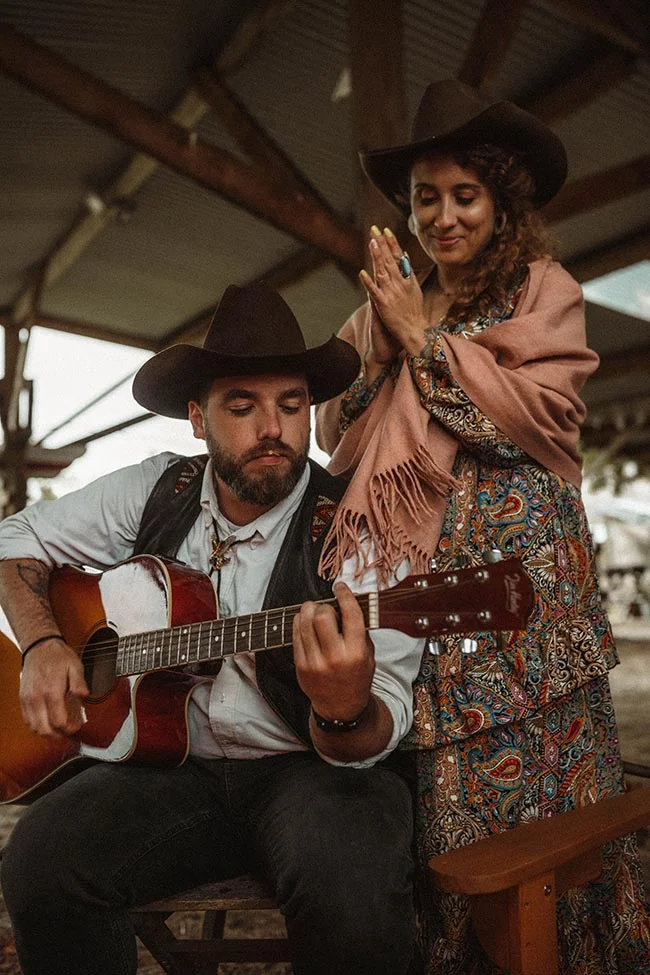 Un couple en tenue western, l'homme jouant de la guitare et la femme souriante, dans une ambiance rustique et chaleureuse.