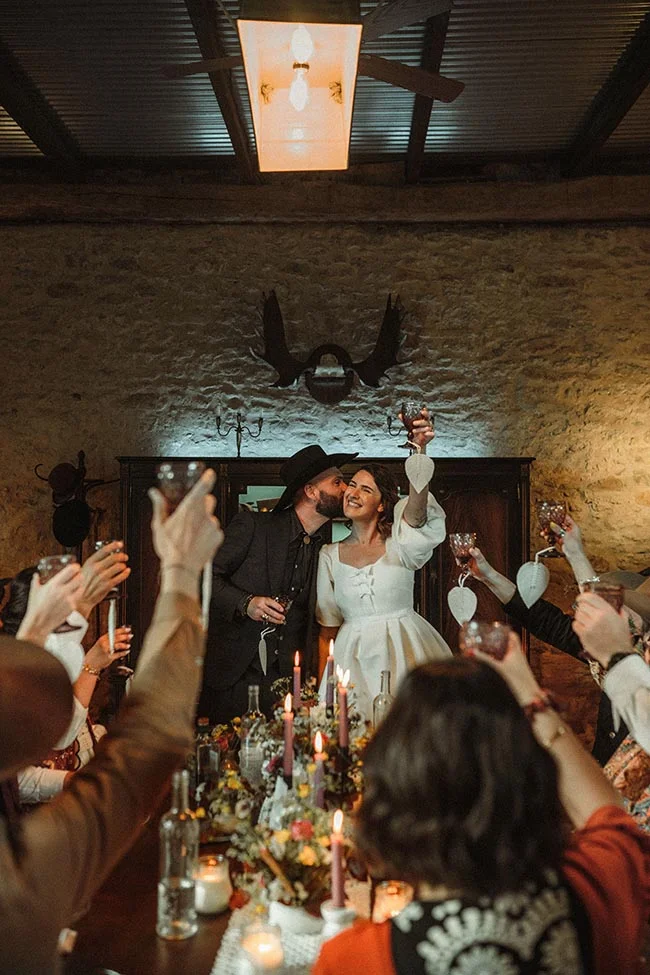 Couple en tenue western trinquant avec des invités lors d'un dîner intime et chaleureux aux tons ocres.