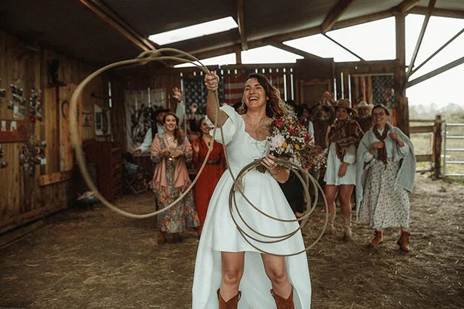 Une mariée joyeuse en robe blanche lance un lasso, entourée d'invités au style western dans une grange rustique.