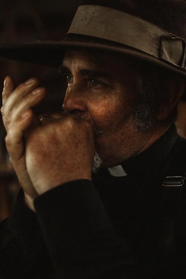 Gros plan d'un prêtre au style western, chapeau brun et col romain, dans une ambiance sombre et intimiste.