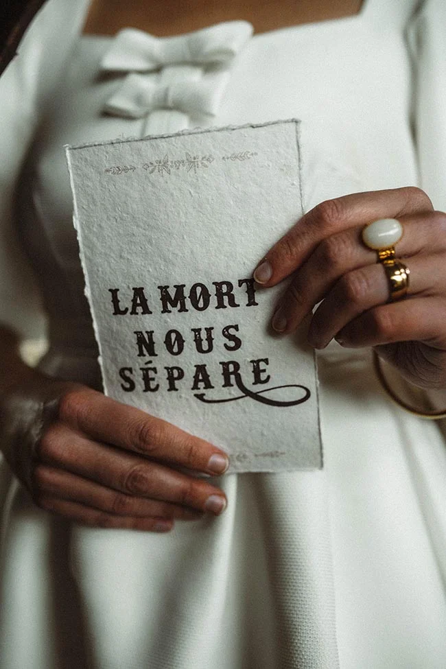 Une mariée en robe blanche tenant un livret blanc avec l'inscription « LA MORT NOUS SÉPARE ».