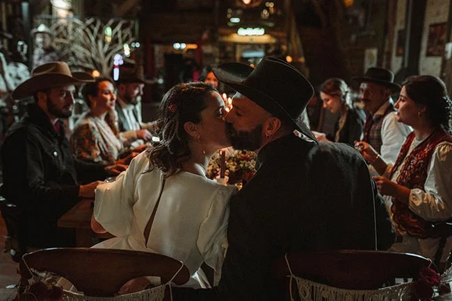 Couple de mariés s'embrassant dans un saloon rustique, entourés d'invités en tenues western dans une ambiance chaleureuse.