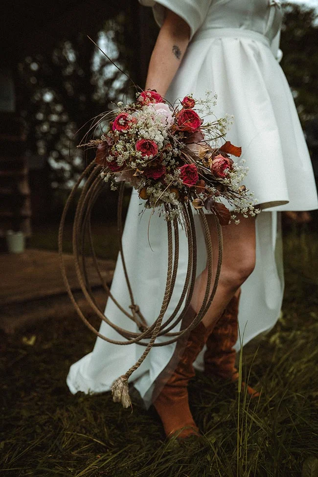Mariée en robe blanche et bottes brunes tenant un bouquet floral fixé à un lasso dans un décor champêtre.