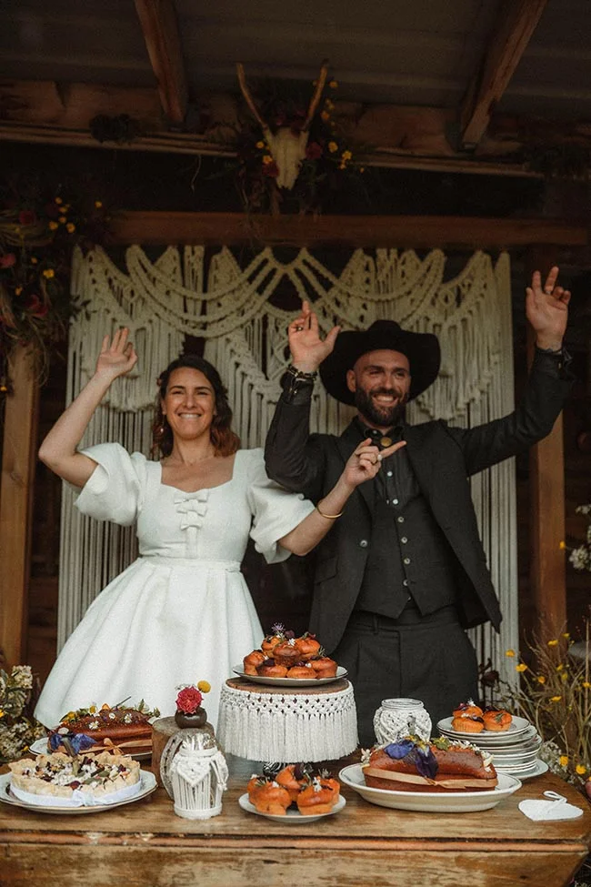 Couple joyeux en tenue western devant un buffet de desserts et un décor macramé bohème.