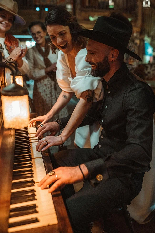 Un couple souriant en tenue western joue du piano dans une ambiance chaleureuse et tamisée.