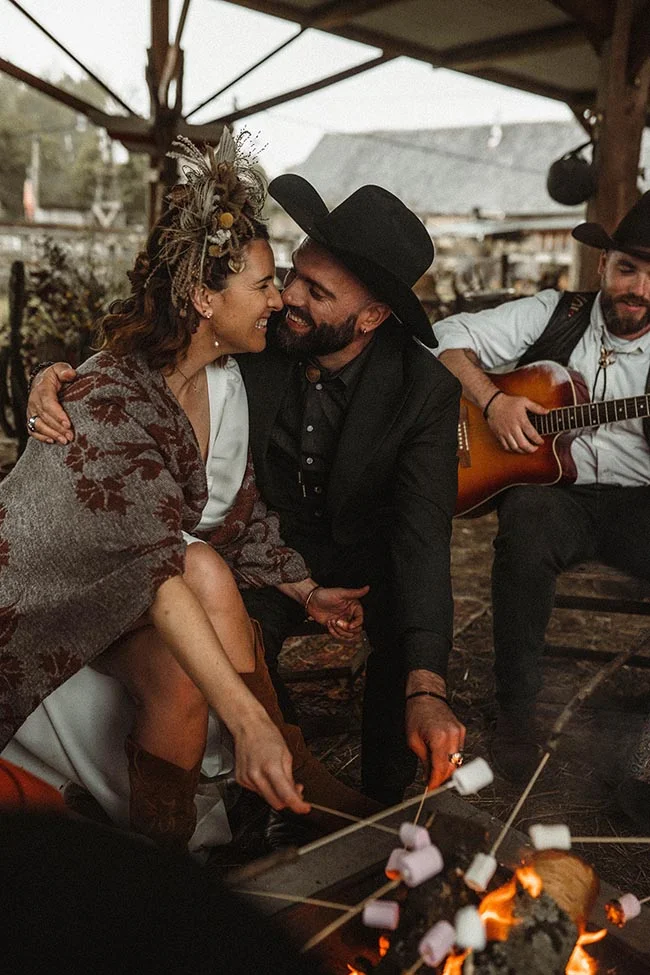 Couple complice en tenue western grillant des guimauves autour d'un feu, ambiance chaleureuse et rustique.