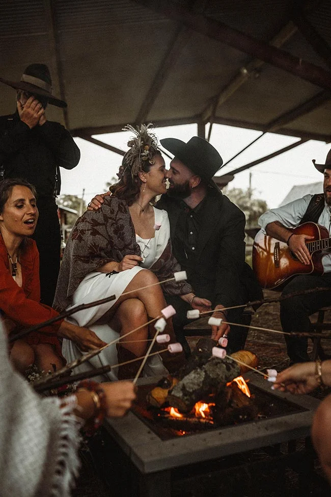 Couple en tenue western s'embrassant près d'un feu de camp entouré d'amis, ambiance chaleureuse et rustique.