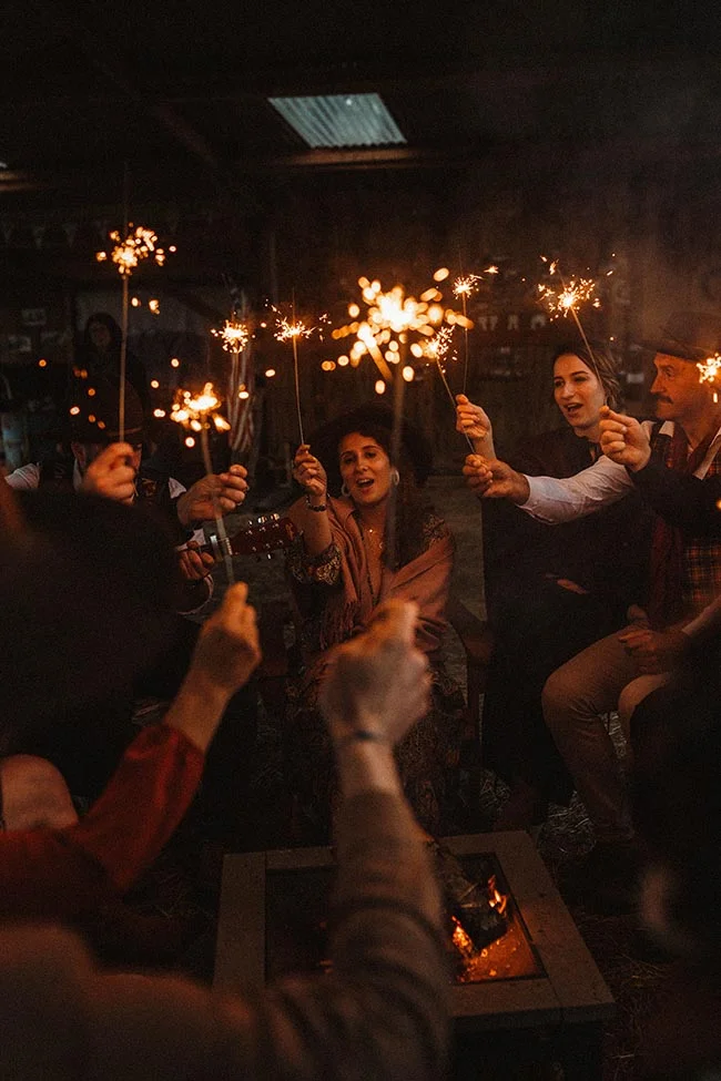 Des invités célèbrent un mariage western avec des cierges magiques autour d'un feu dans une ambiance chaleureuse et sombre.