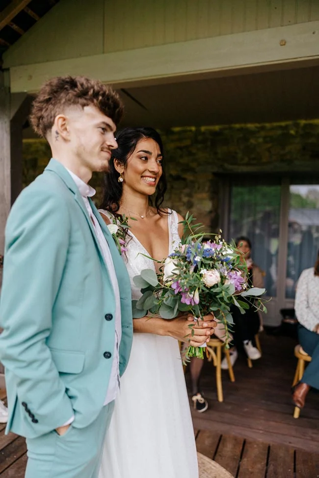 Jeunes mariés souriants, elle en robe blanche avec bouquet champêtre et lui en costume bleu clair, sous un auvent.