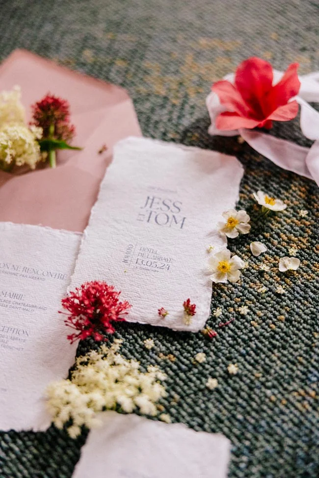 Faire-part de mariage en papier artisanal blanc avec fleurs séchées et fraîches sur un fond texturé sombre.