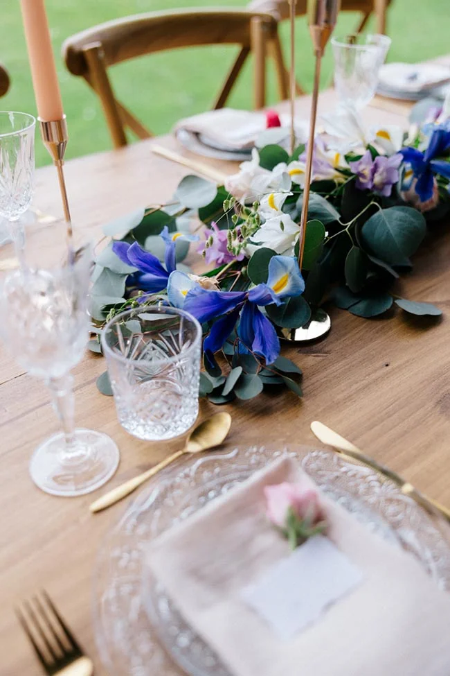 Décoration de table champêtre avec fleurs bleues et blanches, eucalyptus et touches dorées sur bois naturel.