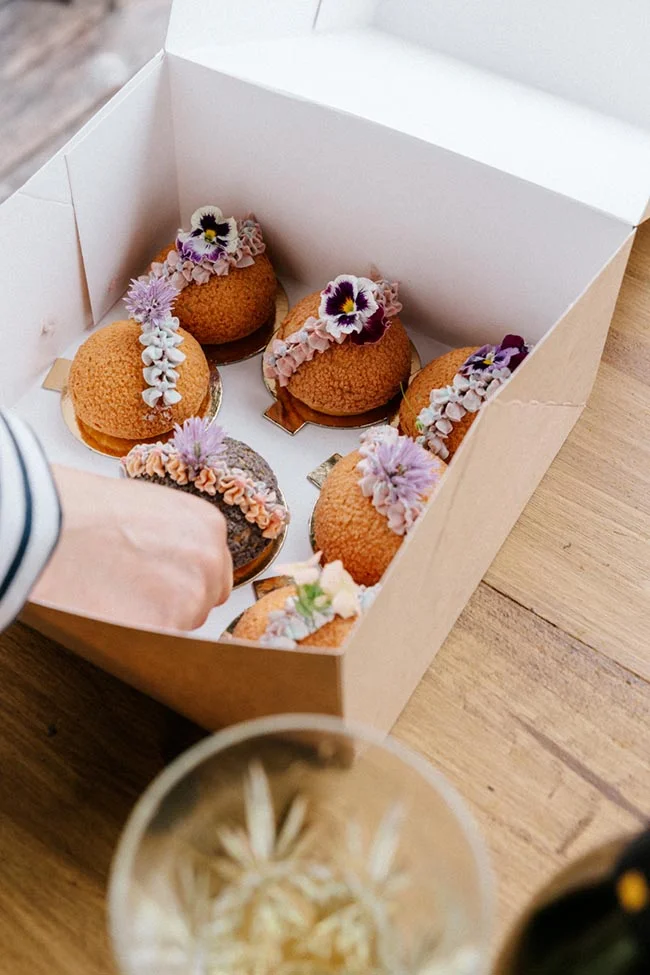 Coffret de pâtisseries artisanales décorées de fleurs comestibles violettes et blanches sur un fond boisé.