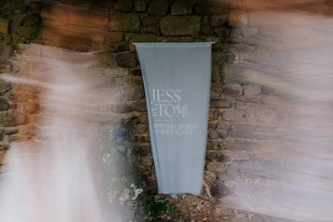 Bannière bleu pâle avec les prénoms Jess et Tomi devant un mur de pierres anciennes, entourée de voiles blancs flous.