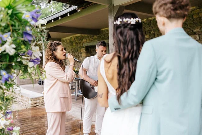 Une chanteuse en costume rose performe pour un couple de mariées sous un auvent en bois, ambiance douce et romantique.