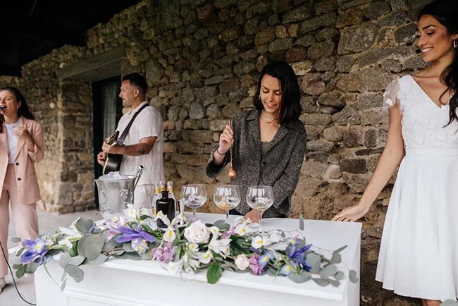 Une femme sert du vin à un buffet fleuri lors d'un mariage convivial devant un mur de pierres rustiques.