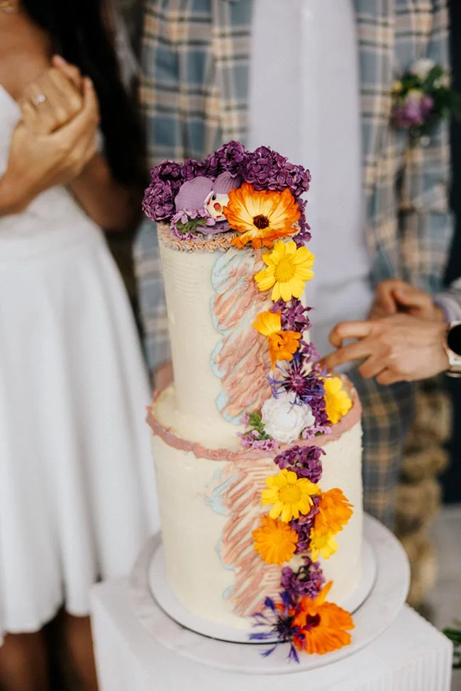 Gâteau de mariage blanc à deux étages, décoré de fleurs colorées et de touches artistiques pastel.