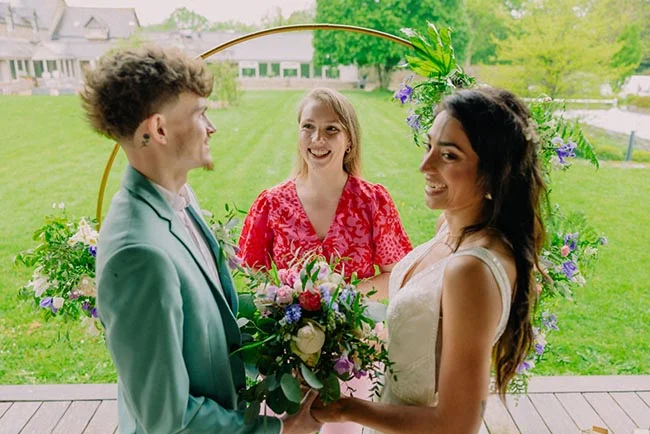 Jeunes mariés et officiante souriants devant une arche fleurie dans un jardin verdoyant.