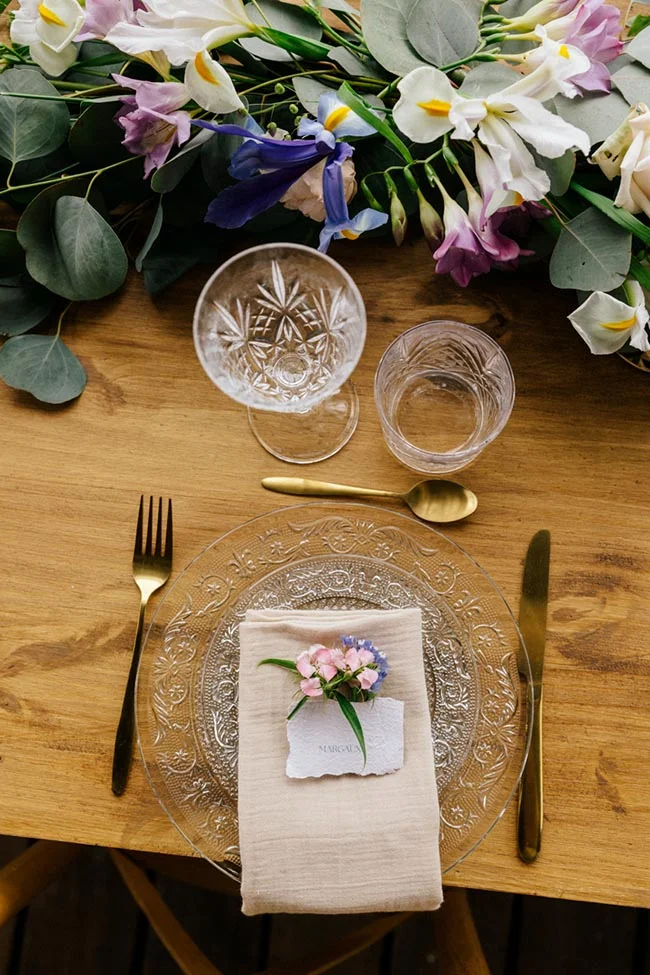 Dressage de table élégant avec assiette en verre, couverts dorés et fleurs printanières sur un bois naturel.