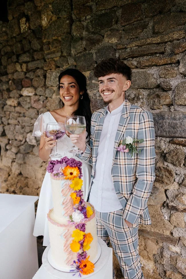Jeunes mariés souriants trinquant devant un gâteau floral coloré, devant un mur de pierres rustiques.