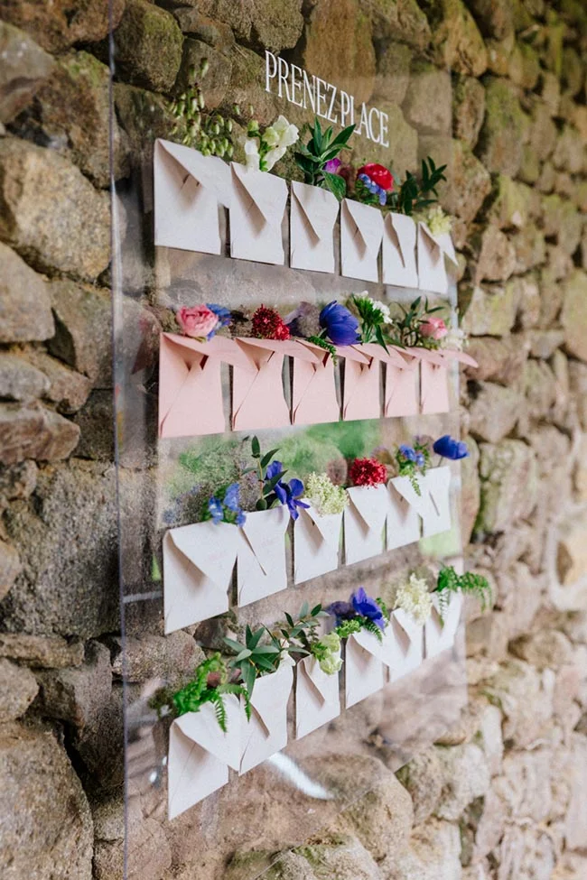 Plan de table minimaliste avec enveloppes roses et blanches et fleurs colorées sur un mur de pierres rustiques.