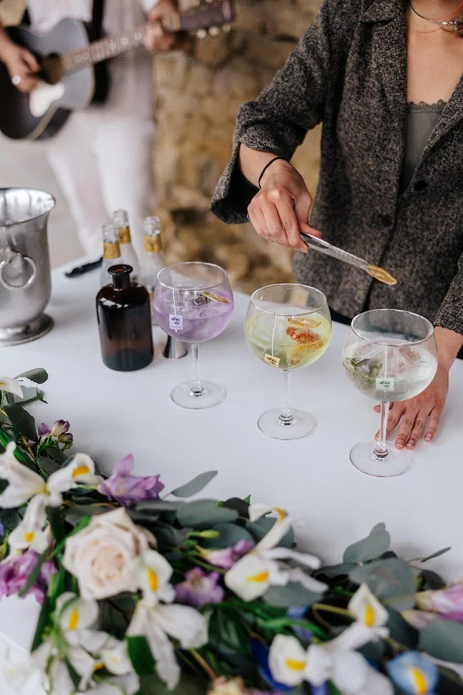 Une main sert des cocktails colorés dans des verres à pied sur une table décorée de fleurs blanches et mauves.
