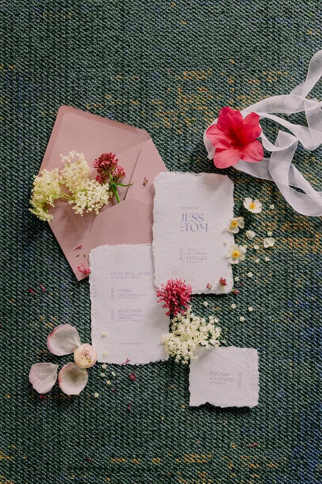 Faire-part de mariage en papier artisanal blanc et enveloppe rose poudré, entourés de fleurs sauvages sur fond texturé.