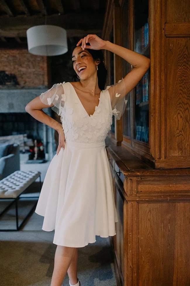 Une mariée souriante en robe blanche courte et vaporeuse, posant avec élégance devant une bibliothèque en bois sombre.