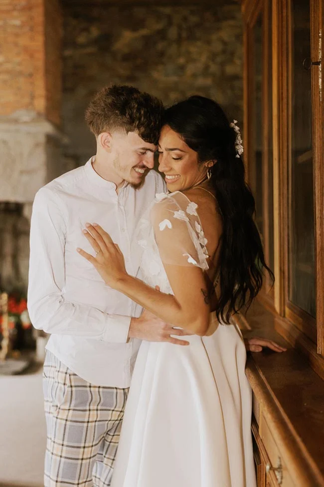 Couple marié complice en tenue blanche, s'enlaçant tendrement dans un intérieur rustique et chaleureux.