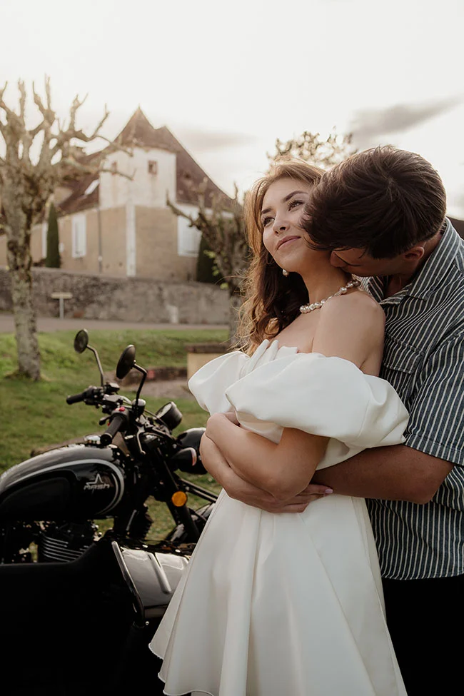 Couple romantique enlacé devant une moto, la mariée en robe blanche, dans un décor champêtre et lumineux.