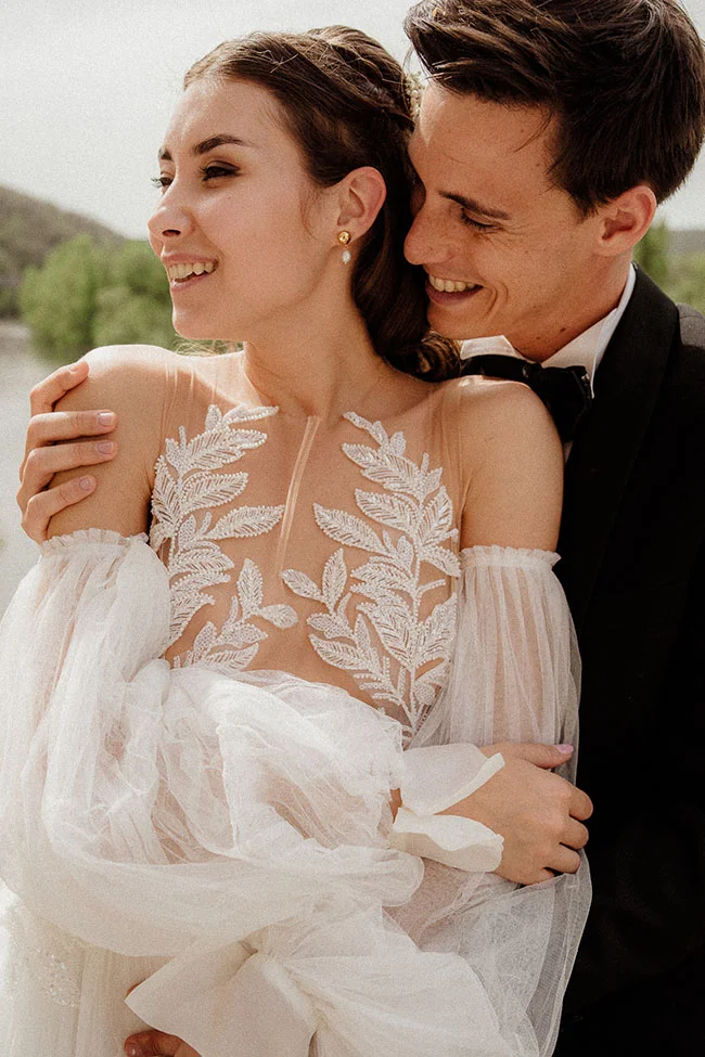 Jeune couple marié souriant et complice, la mariée portant une robe blanche en dentelle et tulle vaporeux.