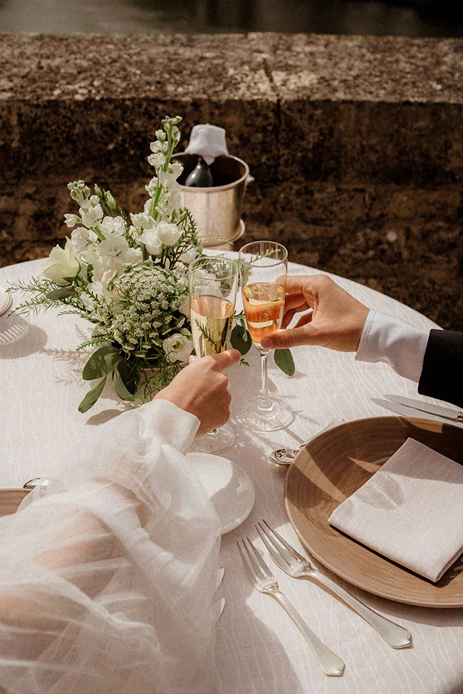 Deux mains trinquant avec des coupes de champagne sur une table élégante avec fleurs blanches et vaisselle beige.
