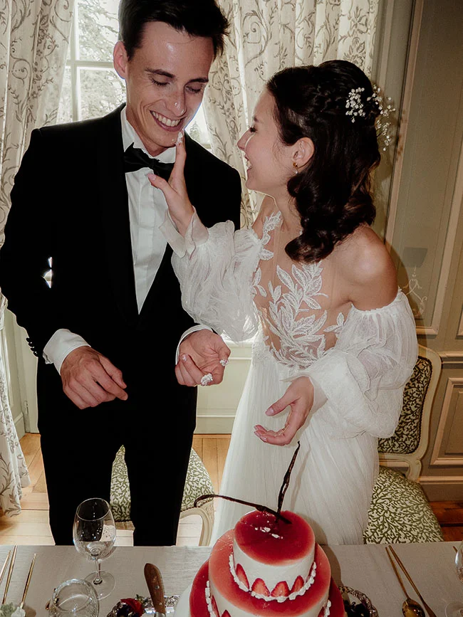 Jeunes mariés complices en tenue formelle blanche et noire, souriant devant un gâteau rouge et blanc.