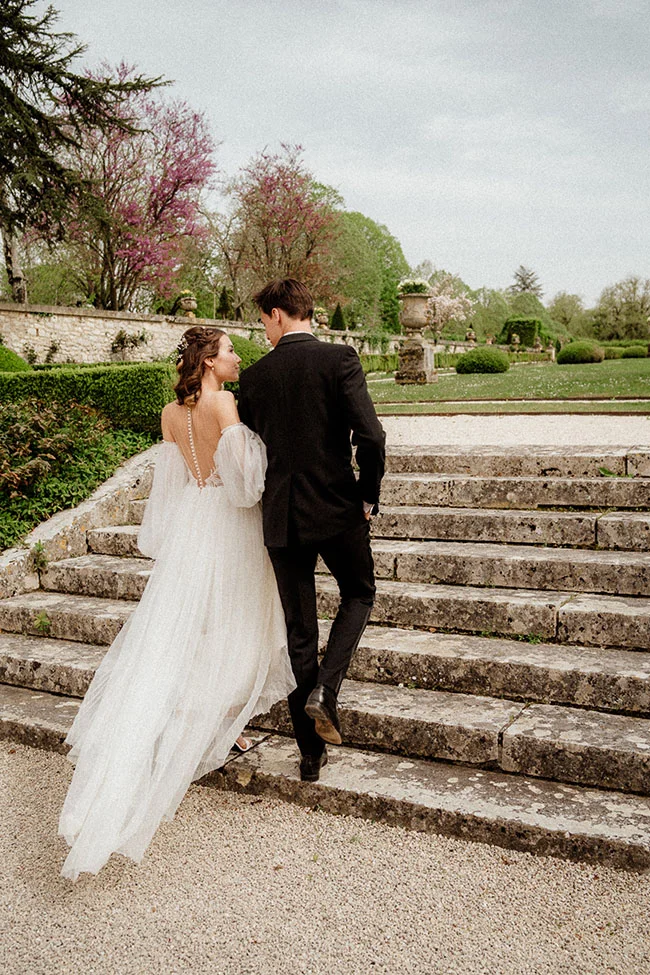 Jeunes mariés en tenue élégante montant des marches en pierre dans un jardin romantique et verdoyant.