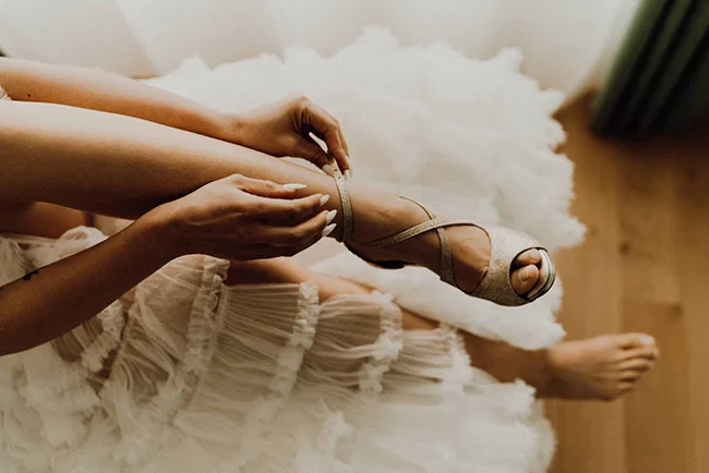Une mariée en robe blanche vaporeuse lace ses sandales argentées dans une ambiance douce et romantique.