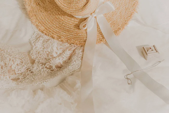 Composition romantique et lumineuse avec un chapeau de paille, une robe en dentelle blanche et une bague de fiançailles.
