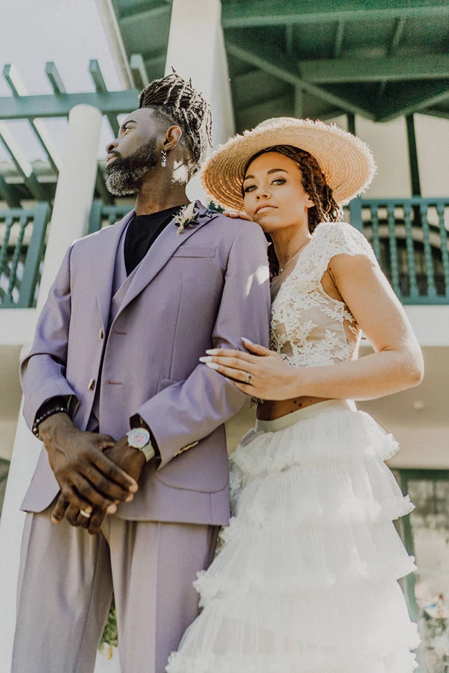 Couple élégant en tenue de mariage mauve et blanche, posant avec romantisme devant une architecture classique.