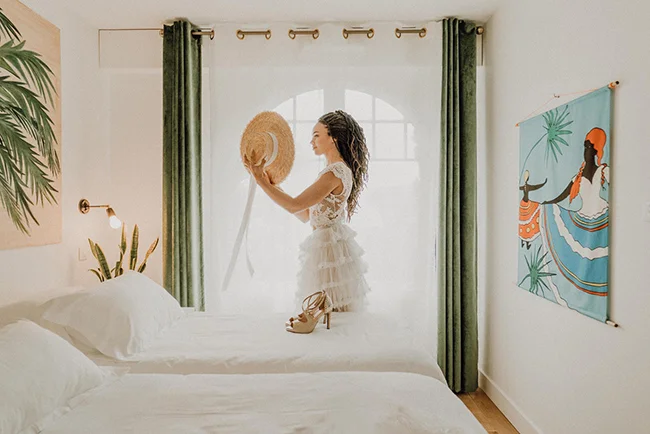 Femme en robe blanche et chapeau de paille dans une chambre lumineuse aux tons vert sauge et blancs.
